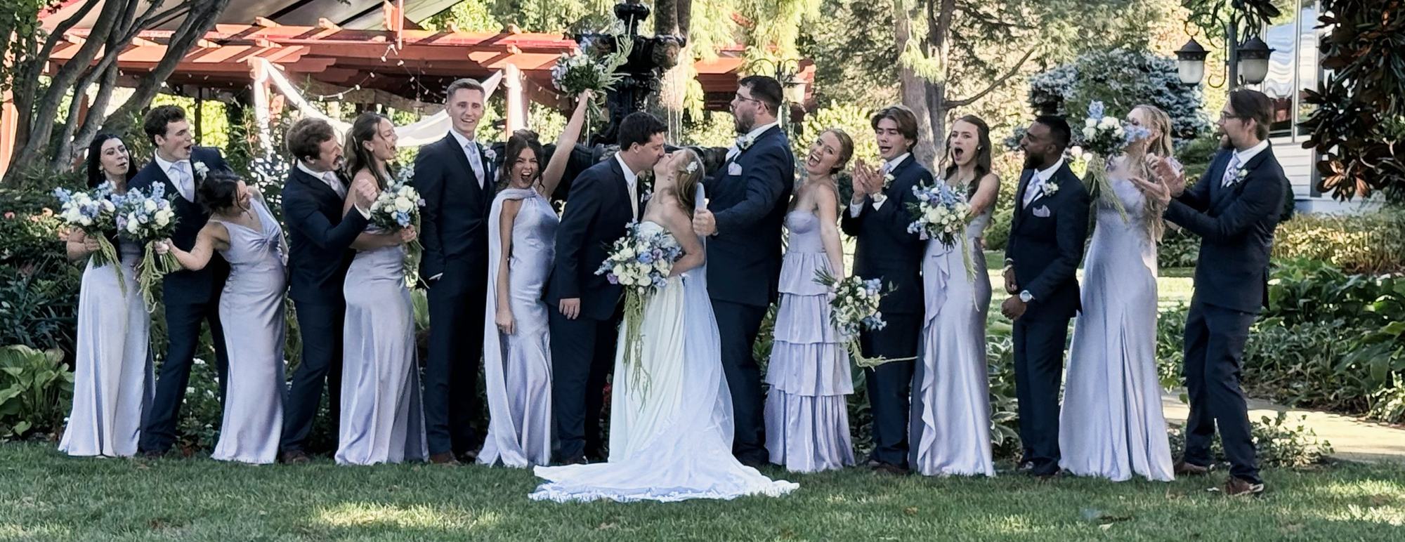 The wedding party in a garden: bridesmaids in lavender dresses, groomsmen in navy suits and the bride in white, laughing together