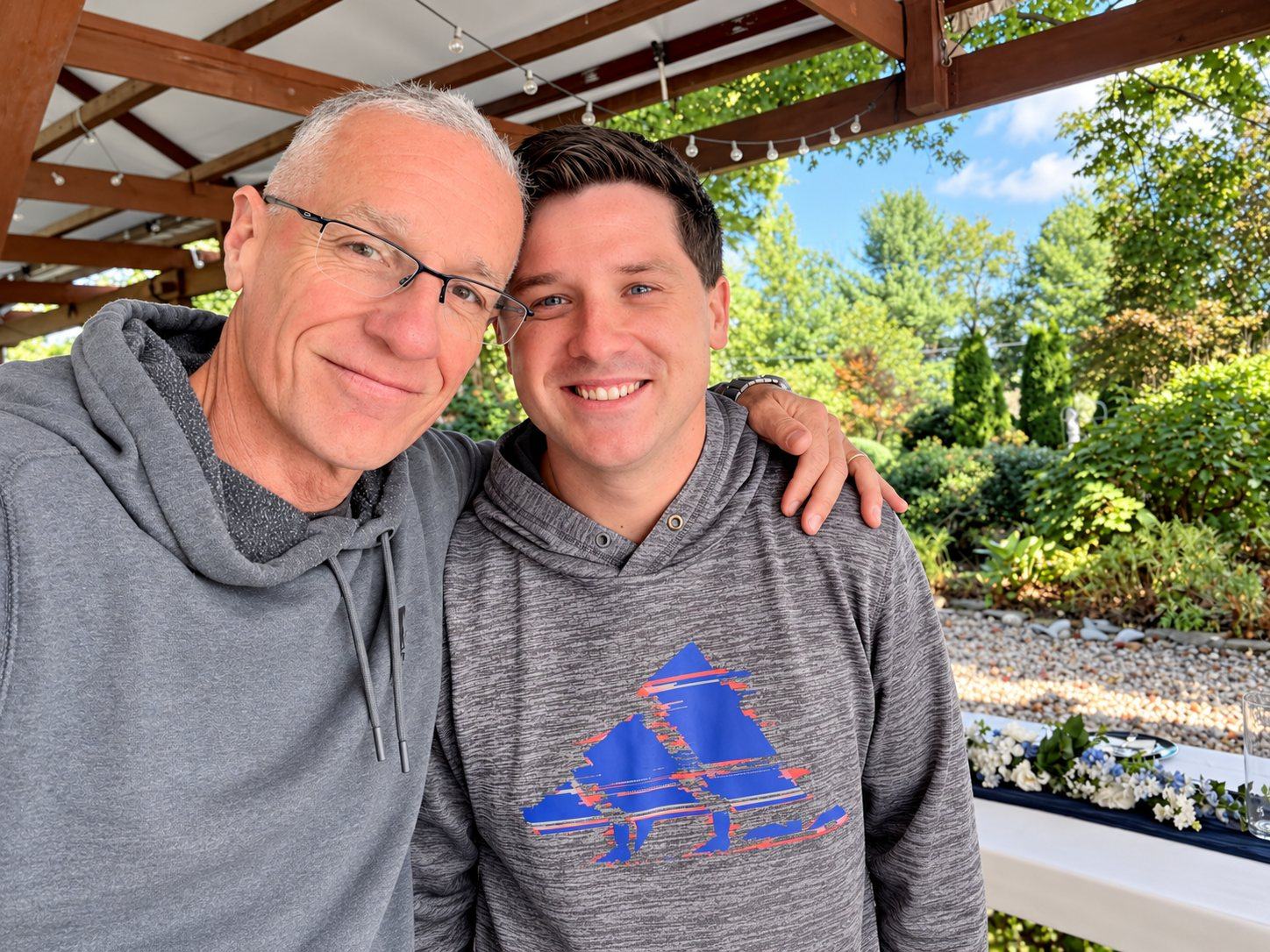 Dan and his son Ryan, cheek to cheek in gray hoodies, smiling under a wooden pergola at the wedding venue