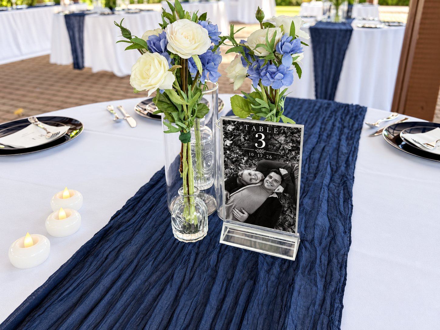 A wedding table set with a navy runner, white and blue flowers, candles and a Table 3 card showing a black-and-white photo of the couple