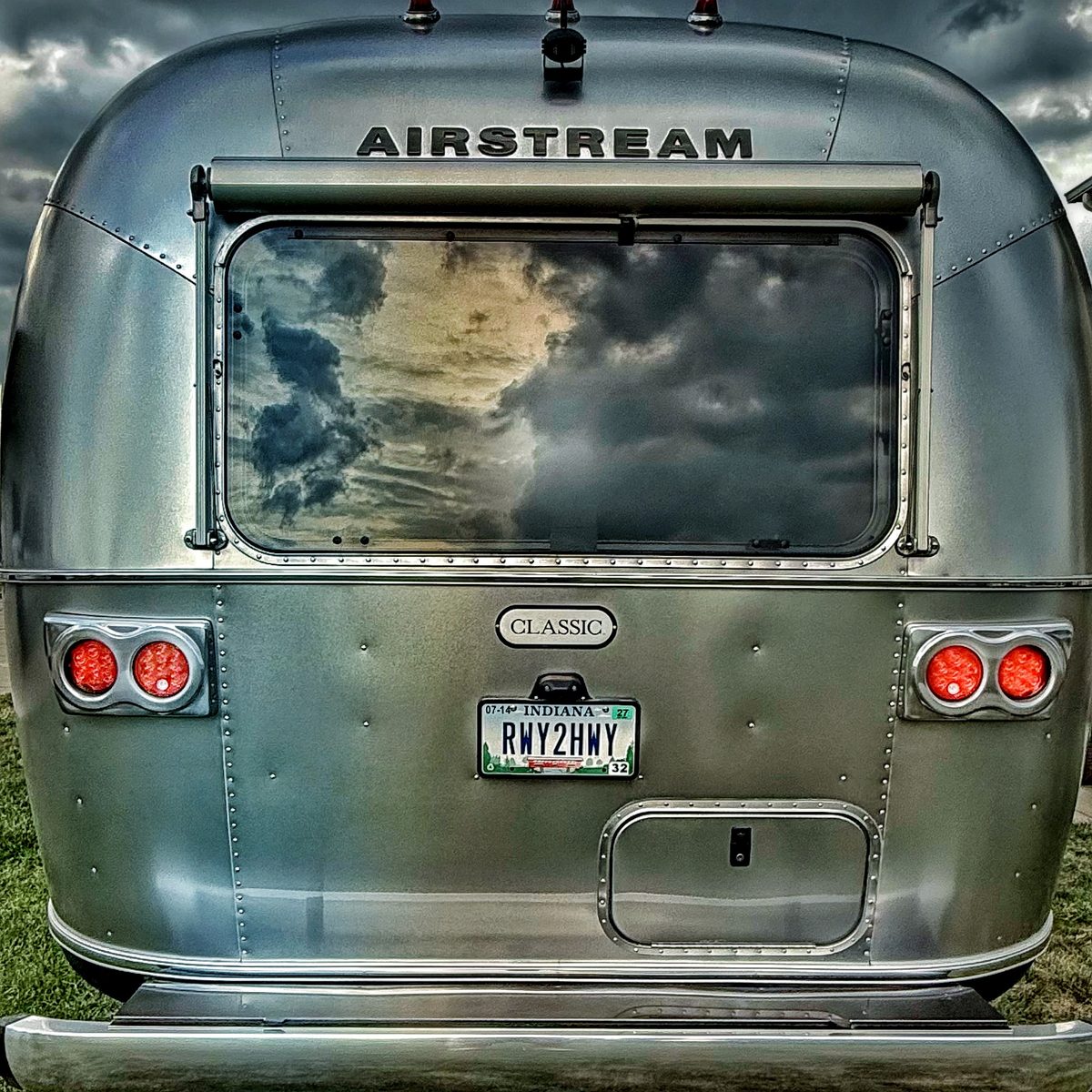 The back of their Airstream Classic under a stormy sky, with an Indiana license plate that reads RWY2HWY