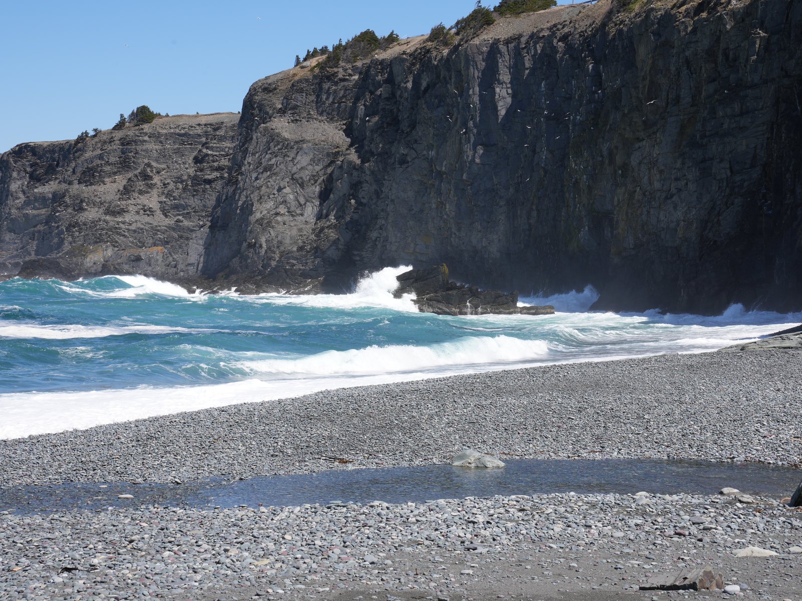 A tall dark cliff wall behind a pebble beach where turquoise waves roll in, with a small pool of seawater on the pebbles