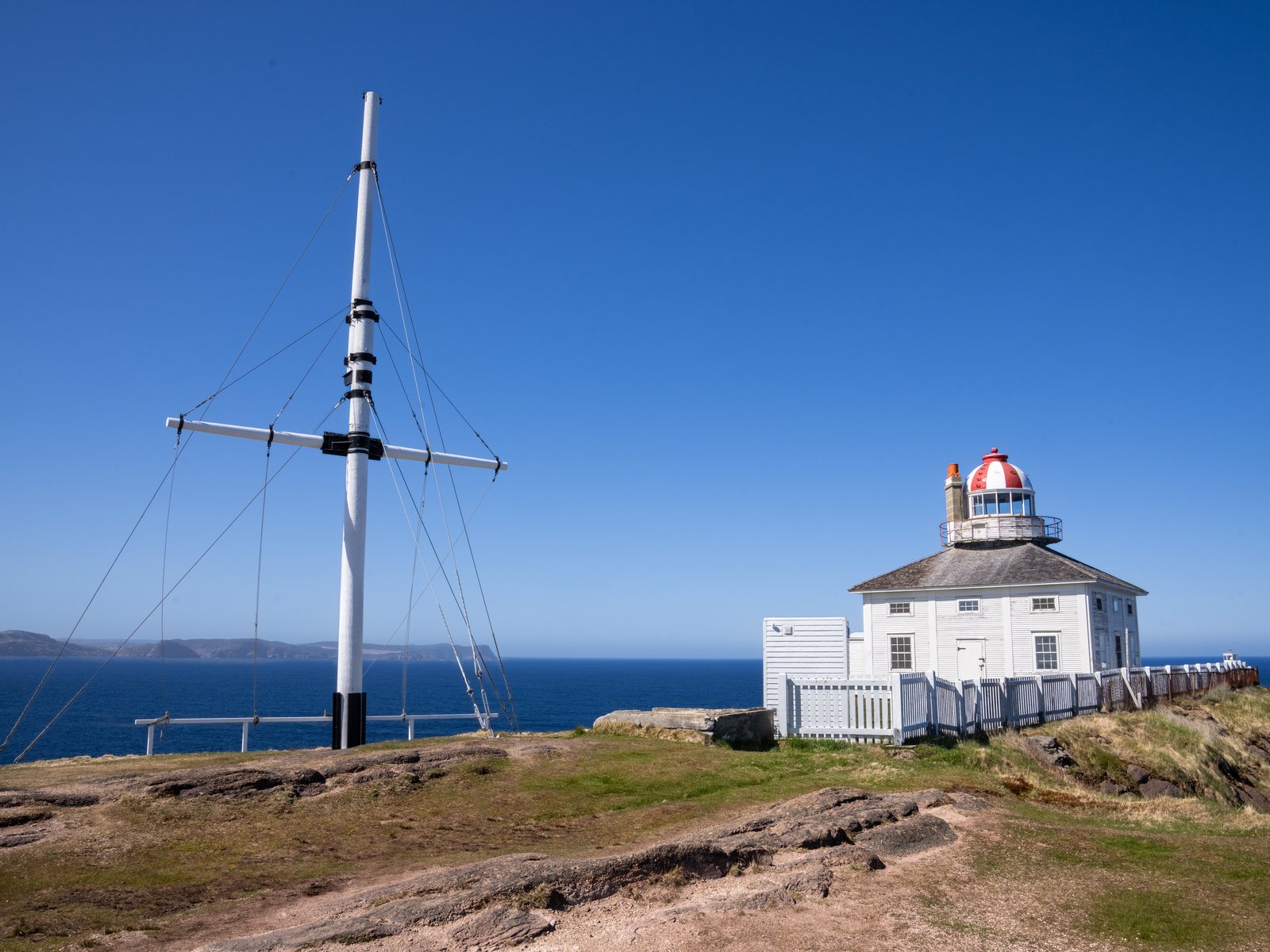 A tall white flagmast with guy wires in front of the old Cape Spear lighthouse on a rocky point