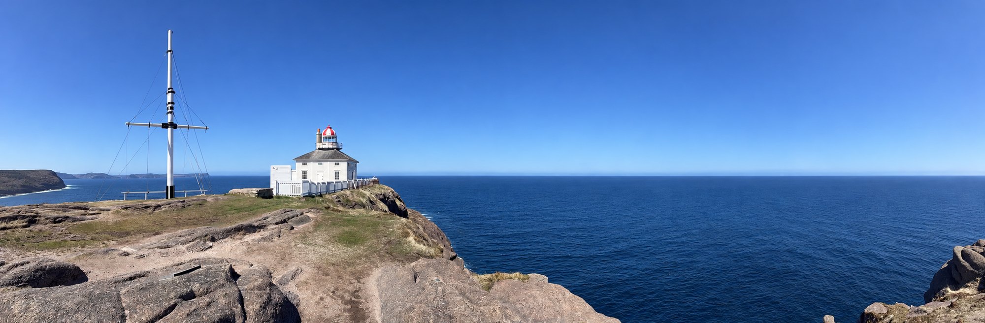 The old Cape Spear lighthouse, a white house with a red-and-white lantern, on a rocky headland beside a tall flagmast, with the ocean behind