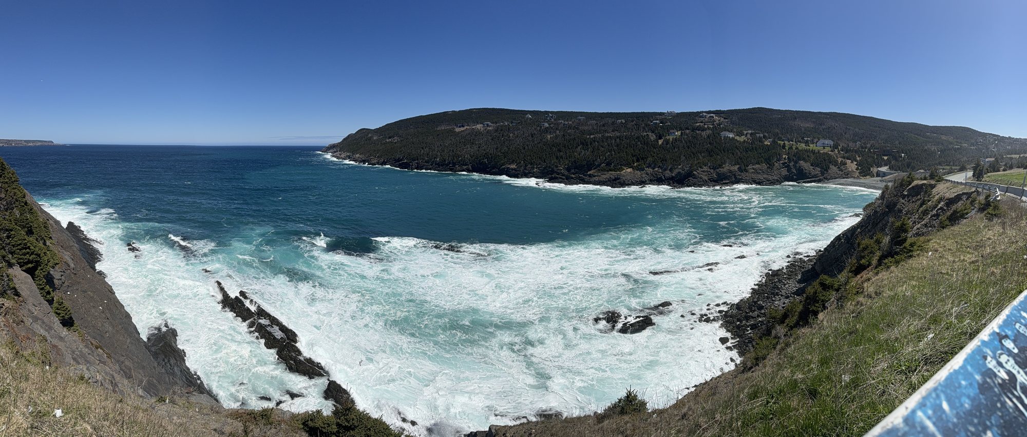 A wide view of a rugged stretch of coast with dark cliffs, white surf at their base and blue sea beyond