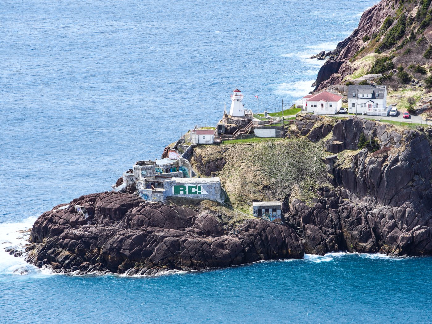 The Fort Amherst lighthouse and white keeper's houses on a dark rocky point, with graffiti-covered concrete fort ruins below