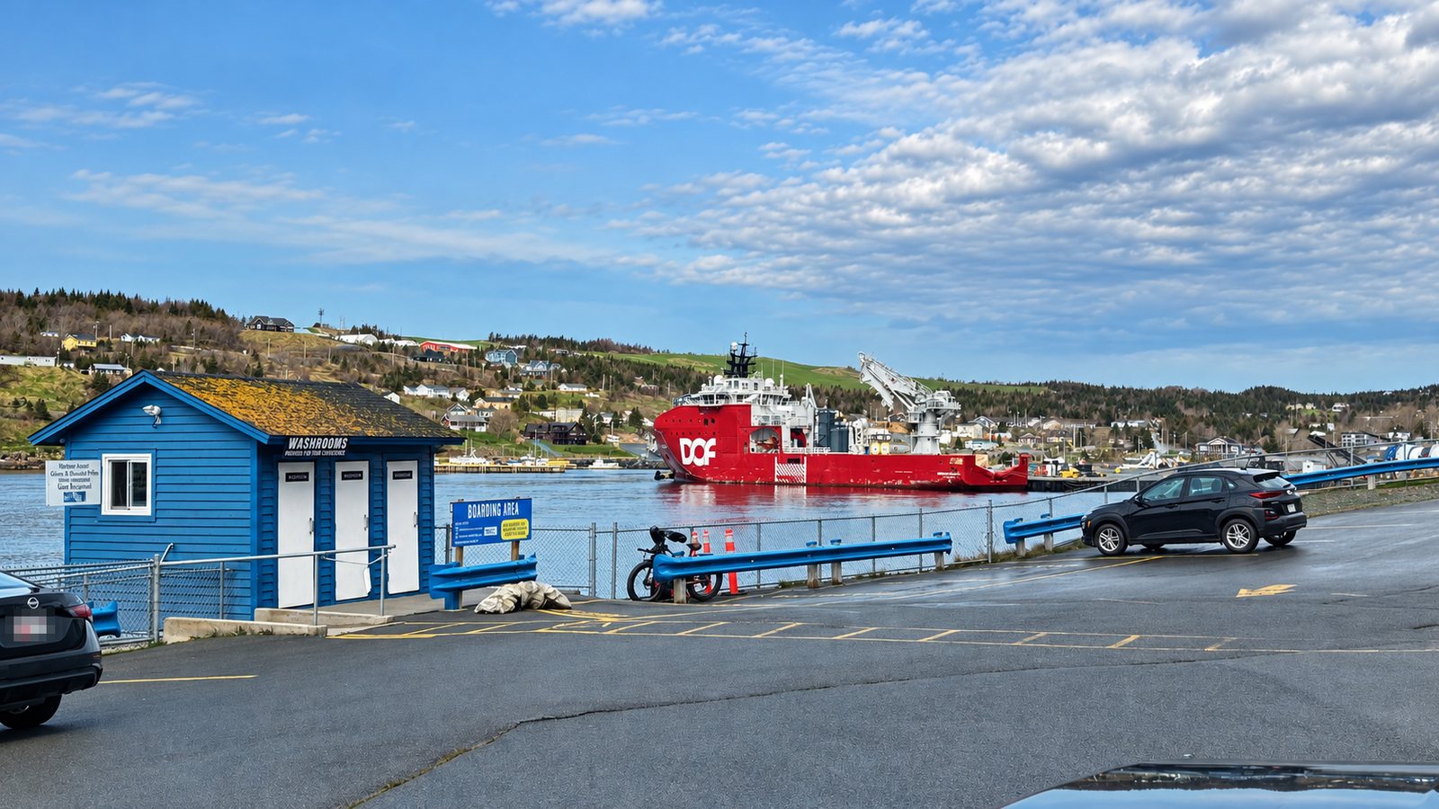 A blue washroom building and a Boarding Area sign beside a paved lot at the water's edge in Bay Bulls, with a red vessel across the harbor