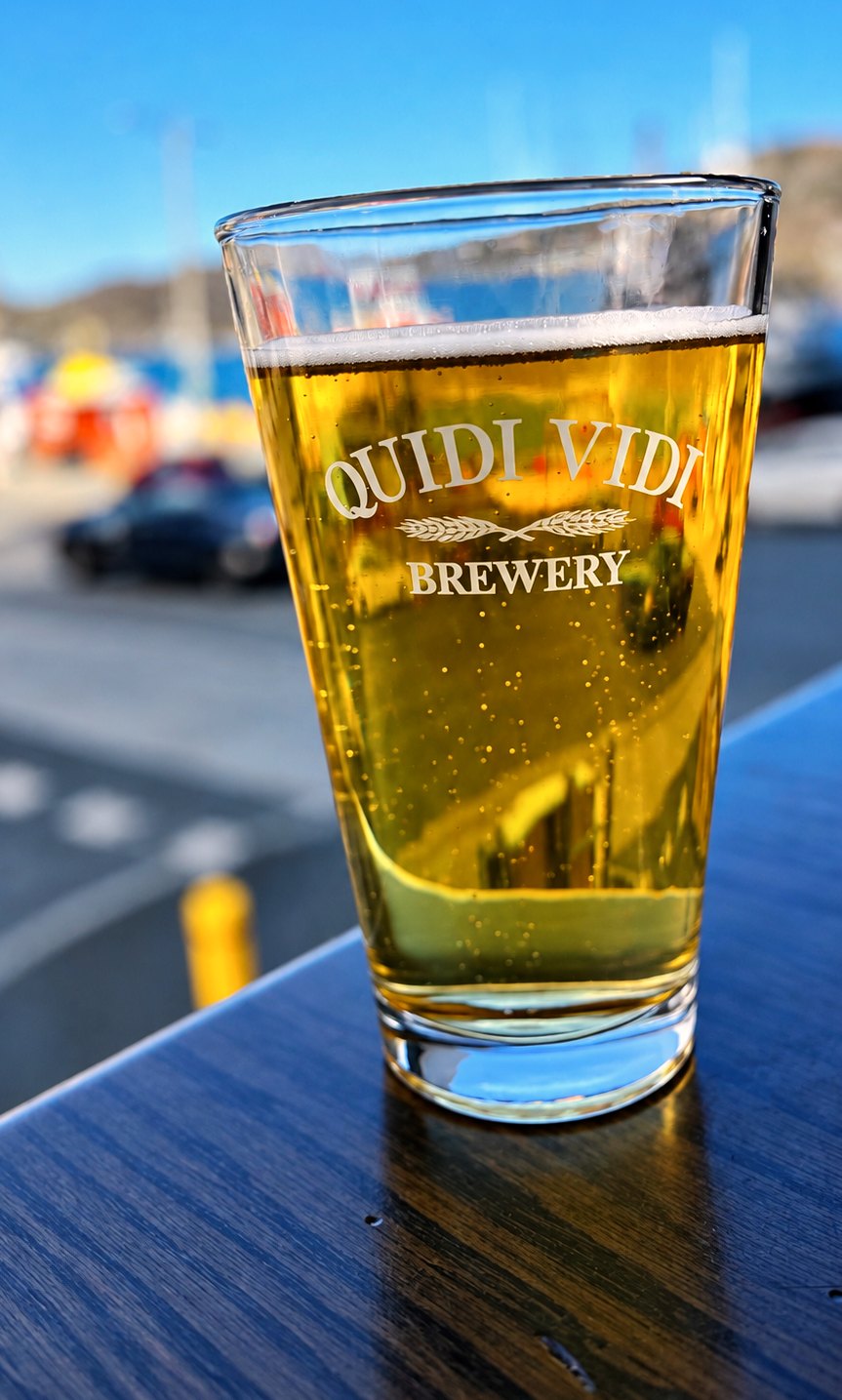 A glass of golden lager printed with Quidi Vidi Brewery on a dark wooden table, with a blurred harbor behind it