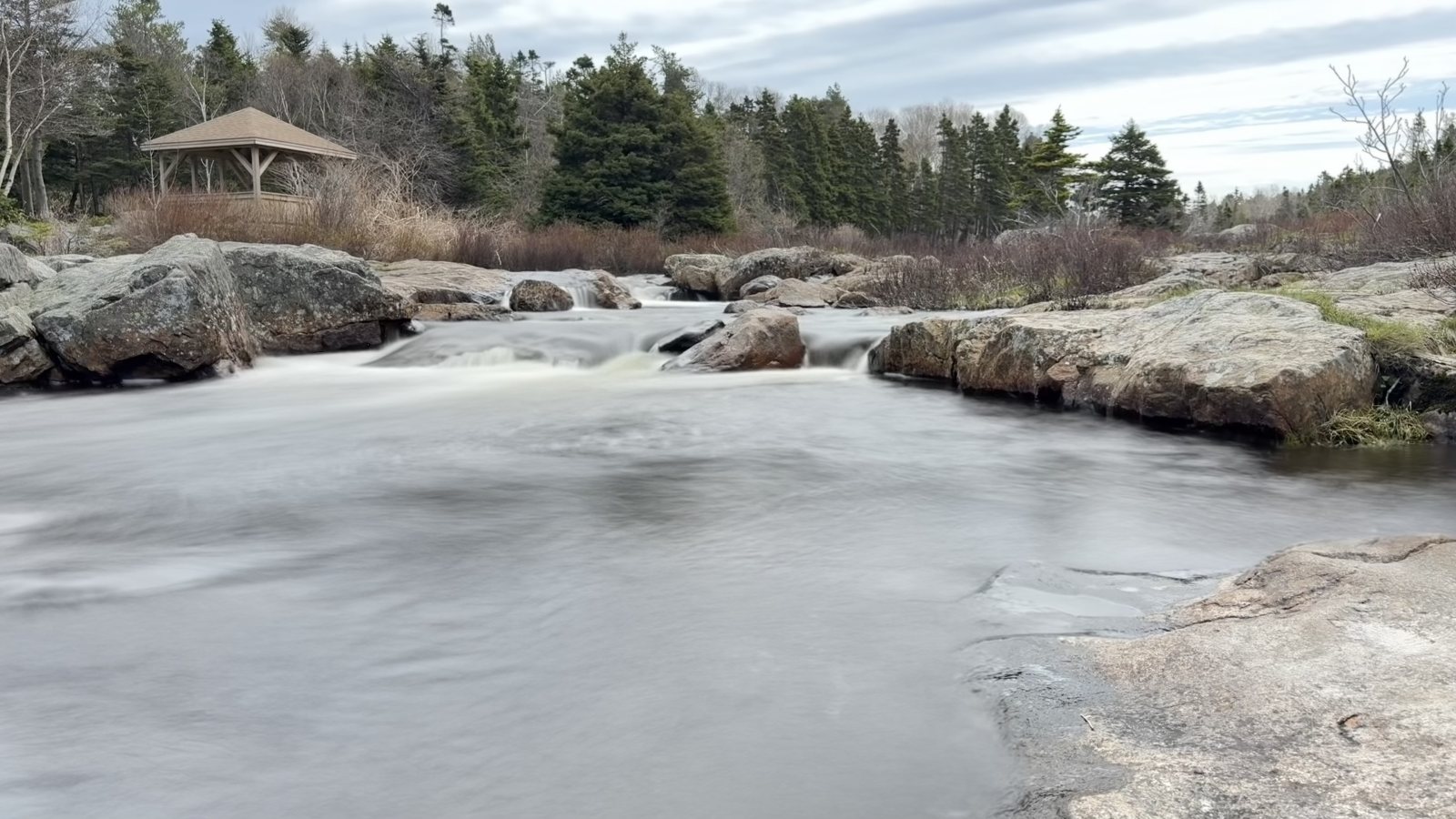 Smooth white water pouring over flat rocks in a river, with a small gazebo and evergreens on the far bank