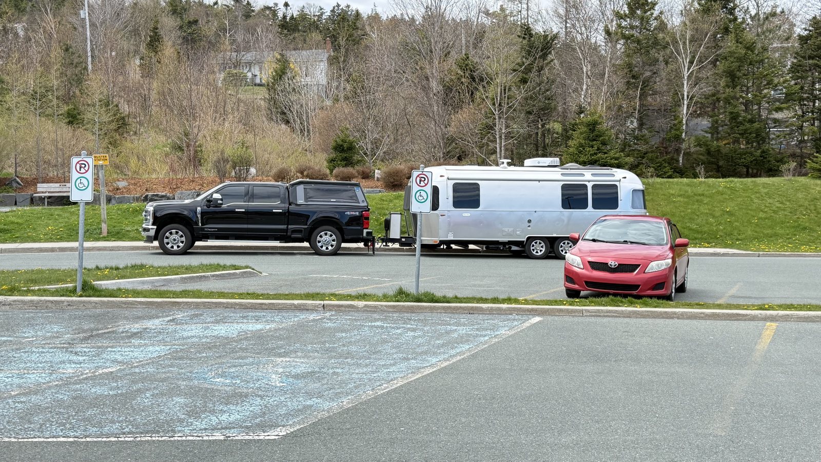 Our Airstream and truck parked in a paved lot beside a grassy area, with a red car, trees and a hillside behind