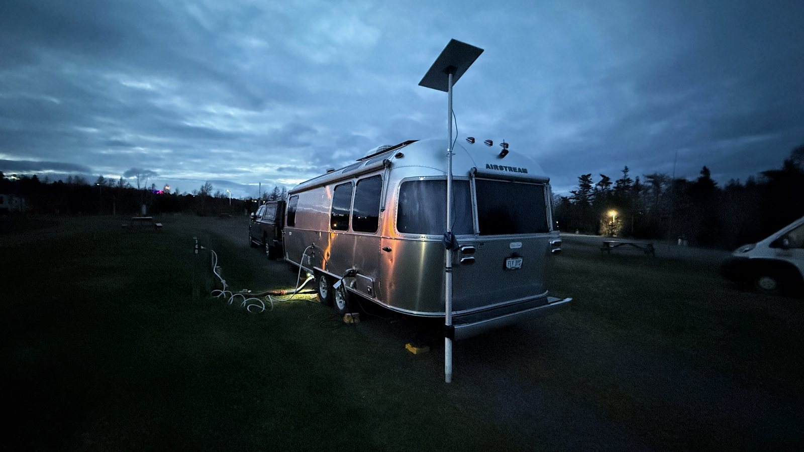 Our Airstream parked on grass at dusk with a Starlink dish on a pole beside it and trees behind