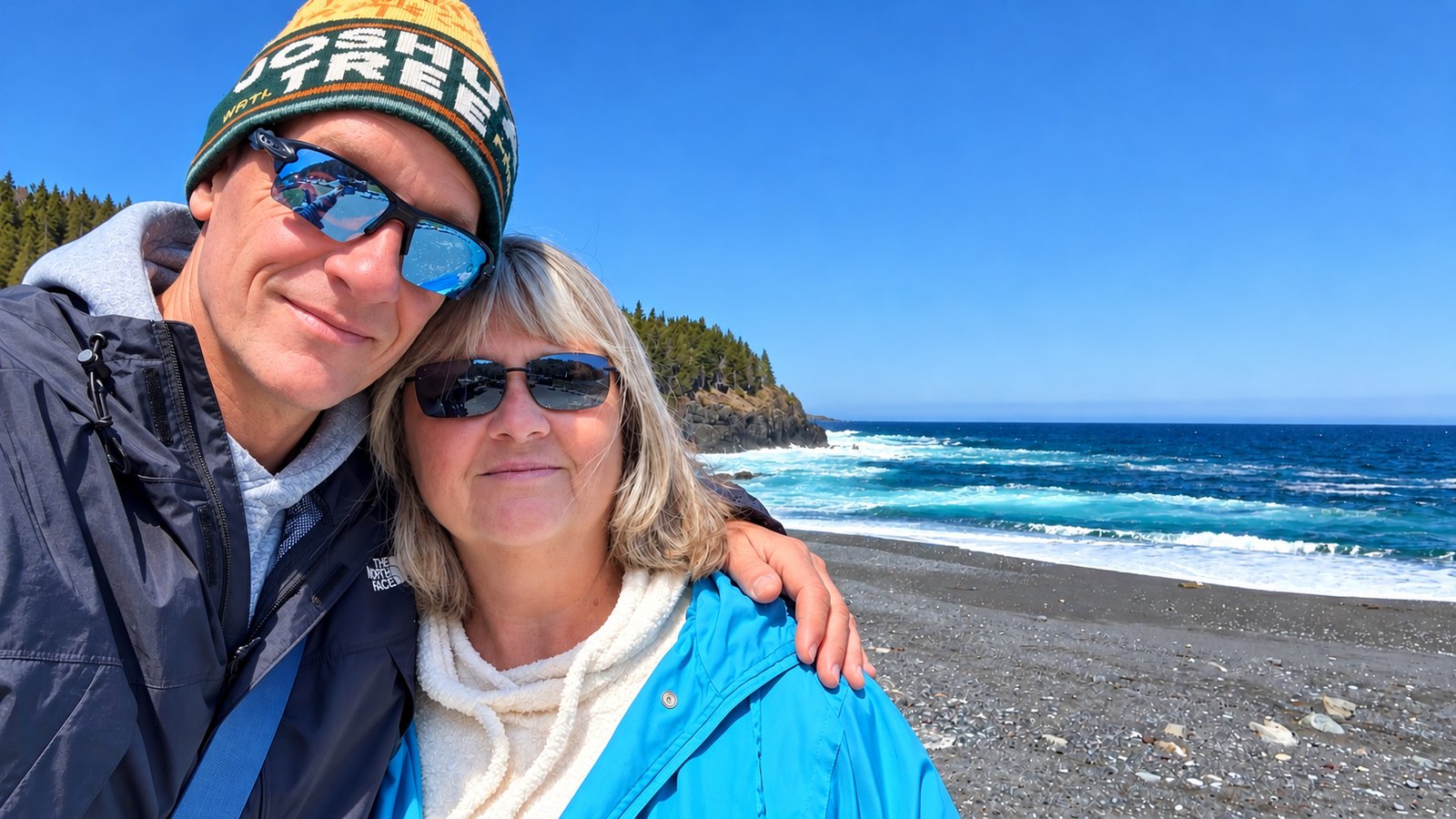 Dan in a knit cap and sunglasses and Heather in sunglasses and a blue jacket, smiling together on a pebble beach with surf behind them