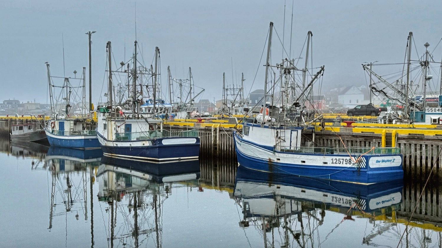 Blue and white fishing boats tied up along a yellow-edged dock, their masts reflected in still water