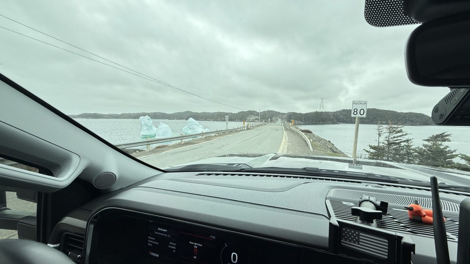 View through the truck windshield of a causeway road running across the water, with pale-blue ice beside the guardrail