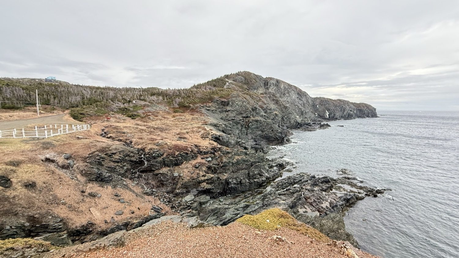 A rocky headland dropping to gray water at Crow Head, with a white fence and a gravel road at the left