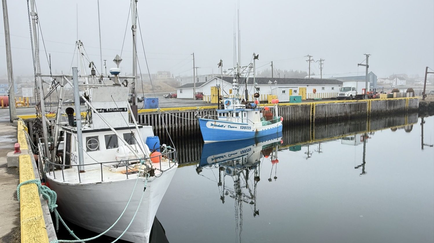 Fishing boats moored beside a dock, with warehouses and utility poles fading into fog