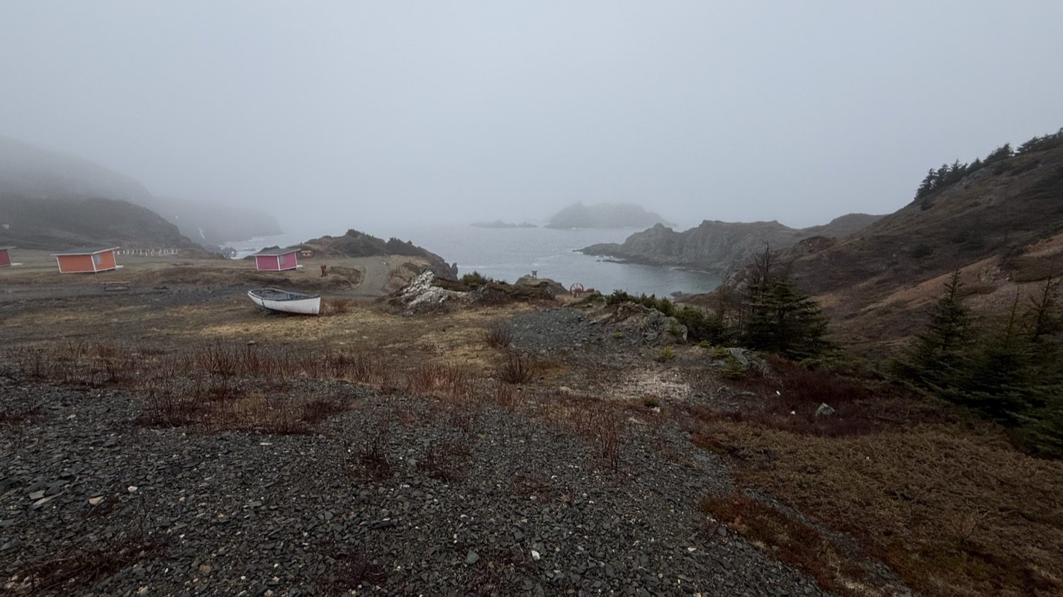 A white boat resting on the shore of a small cove, with red and orange sheds and fog-covered hills behind it