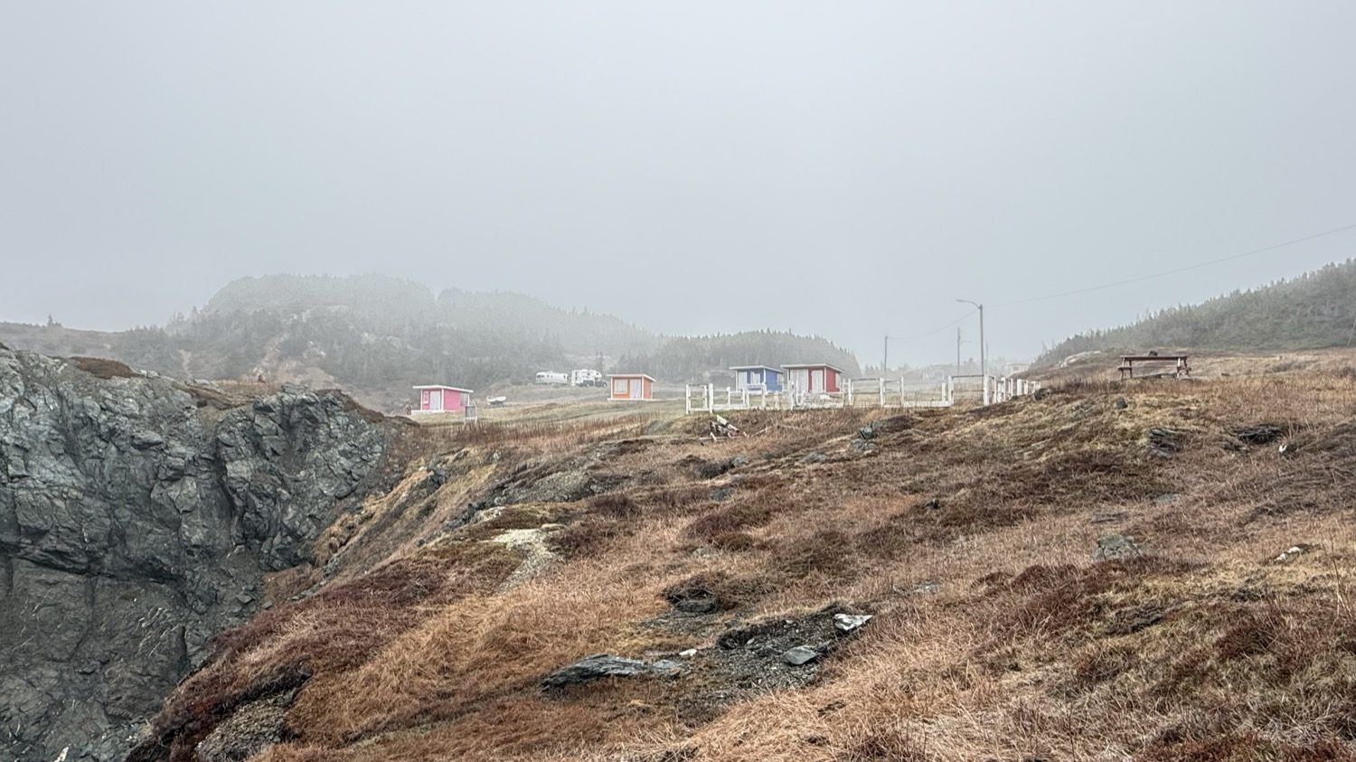 Small colorful sheds and a fenced walkway on a grassy hillside above the cove, with fog covering the hills behind