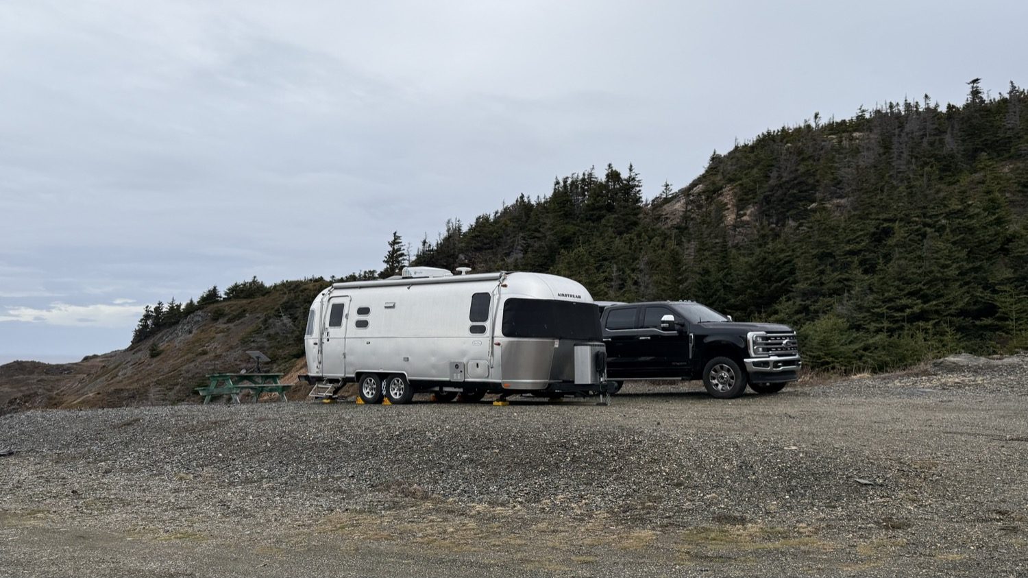 Our truck and Airstream parked on a gravel lot beside a green picnic table, with a tree-covered hill behind
