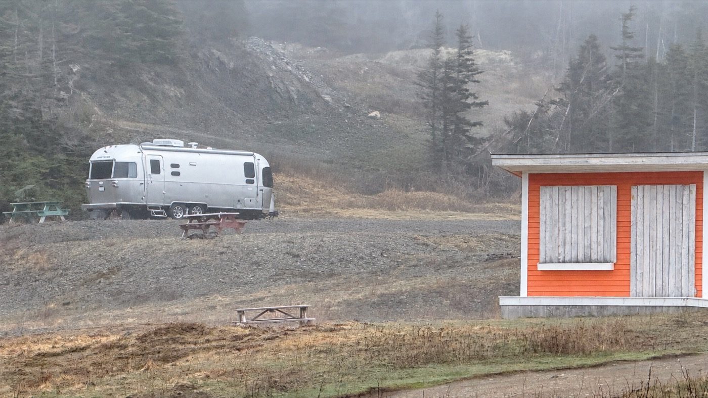 The Airstream parked on the gravel lot in fog, beside an orange shed