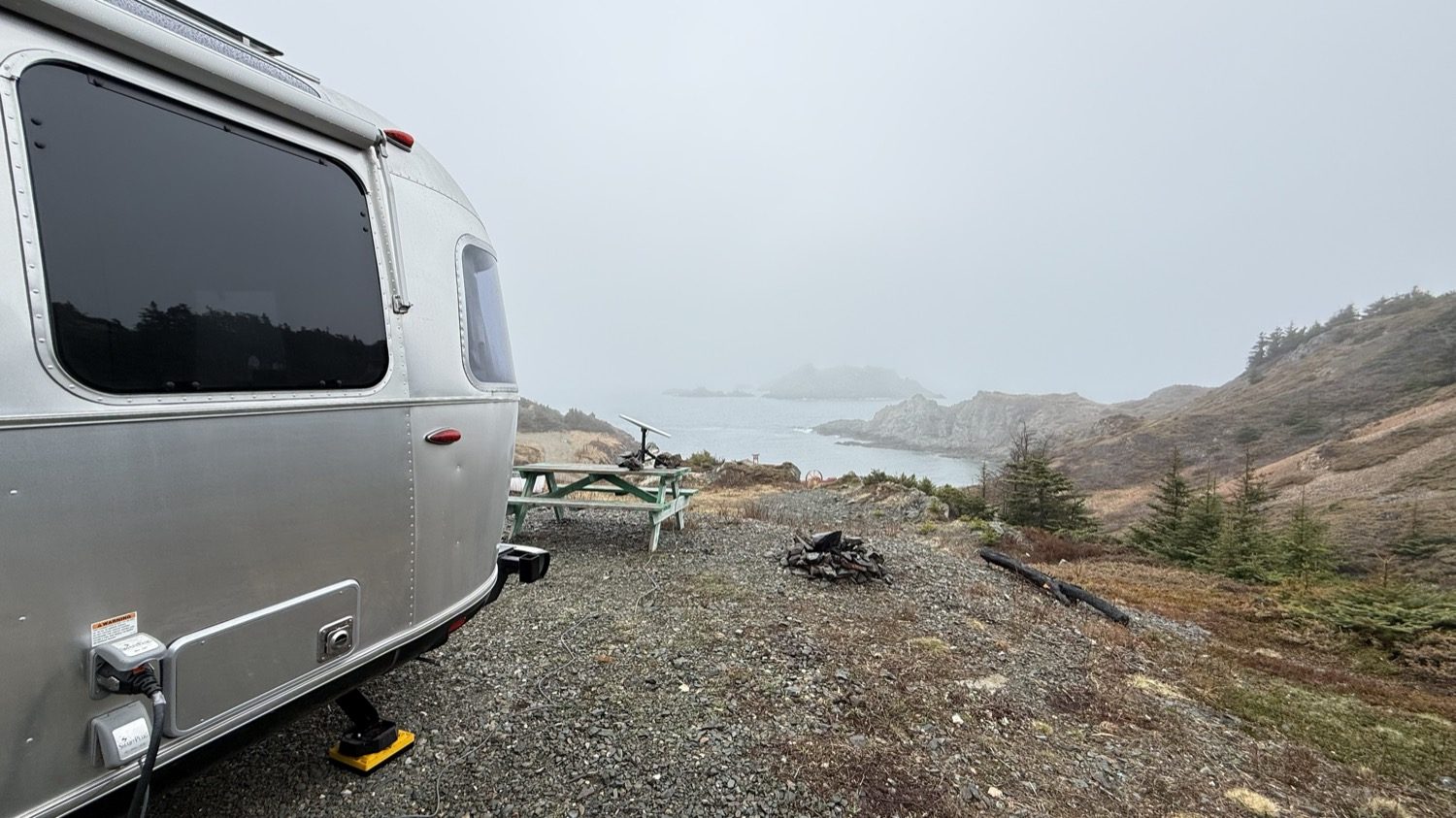The back of our Airstream beside a picnic table and fire ring, with a foggy cove beyond
