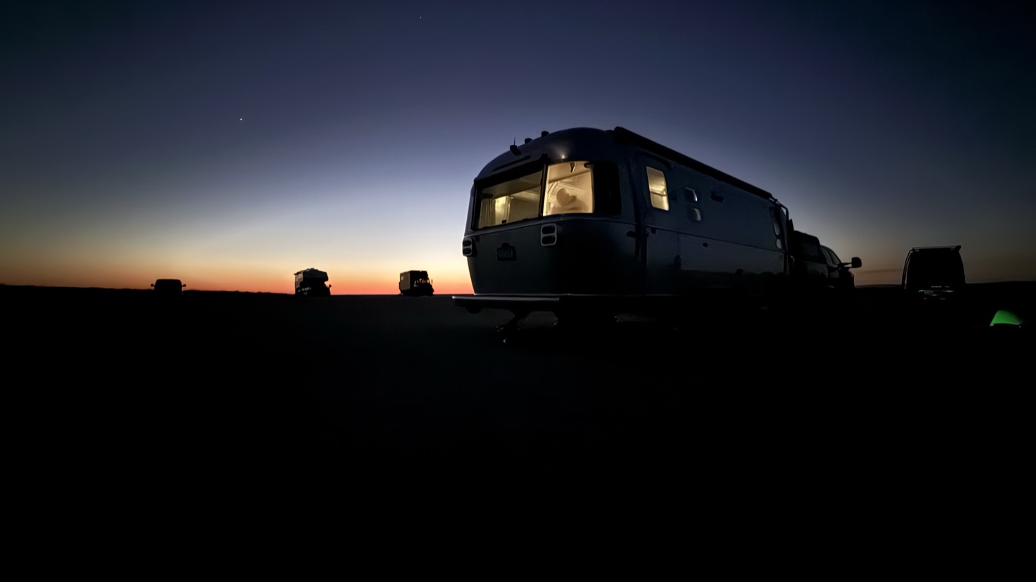 The Airstream glowing from within against a darkening blue and orange sky at Wreckhouse, with other campers silhouetted in the lot