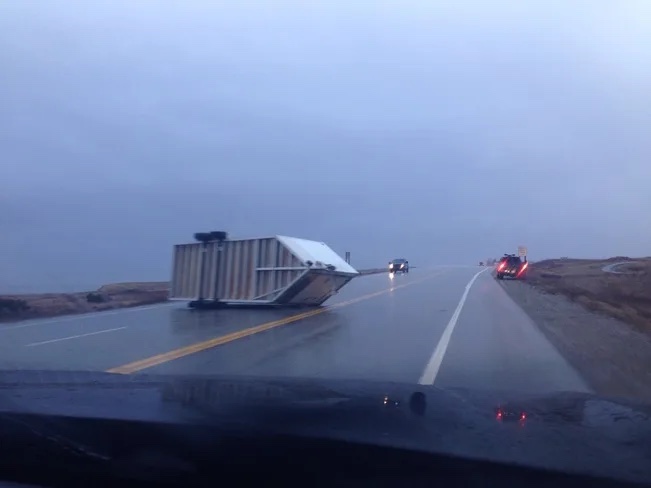 A large trailer lying on its side across the lane of a wet highway at Wreckhouse after a windstorm, with vehicles stopped nearby